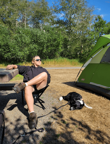 Hal sitting in a camping chair next to a small black dog, the two of them are flanked by a wooden picnic table to the left and a camping tent to the right.