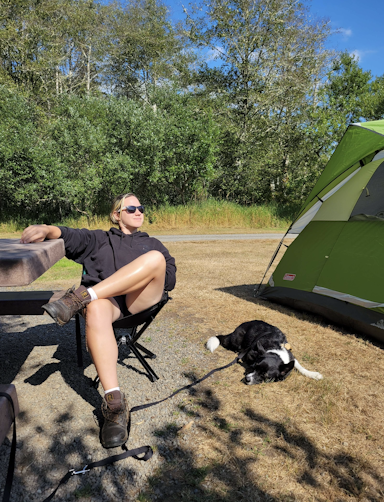 Hal sitting in a camping chair next to a small black dog, the two of them are flanked by a wooden picnic table to the left and a camping tent to the right.